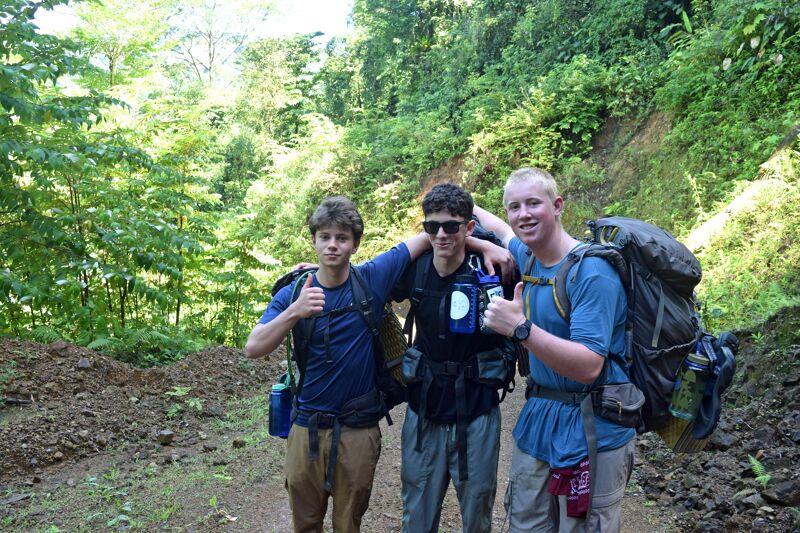 Three young men are posing for a photo on a trail. They are in a wooded area, possibly hiking, as they have backpacks. The man in the center is wearing sunglasses. All three are giving a thumbs-up. The trail appears to be muddy or dirt-covered. The background is filled with lush green trees and vegetation.